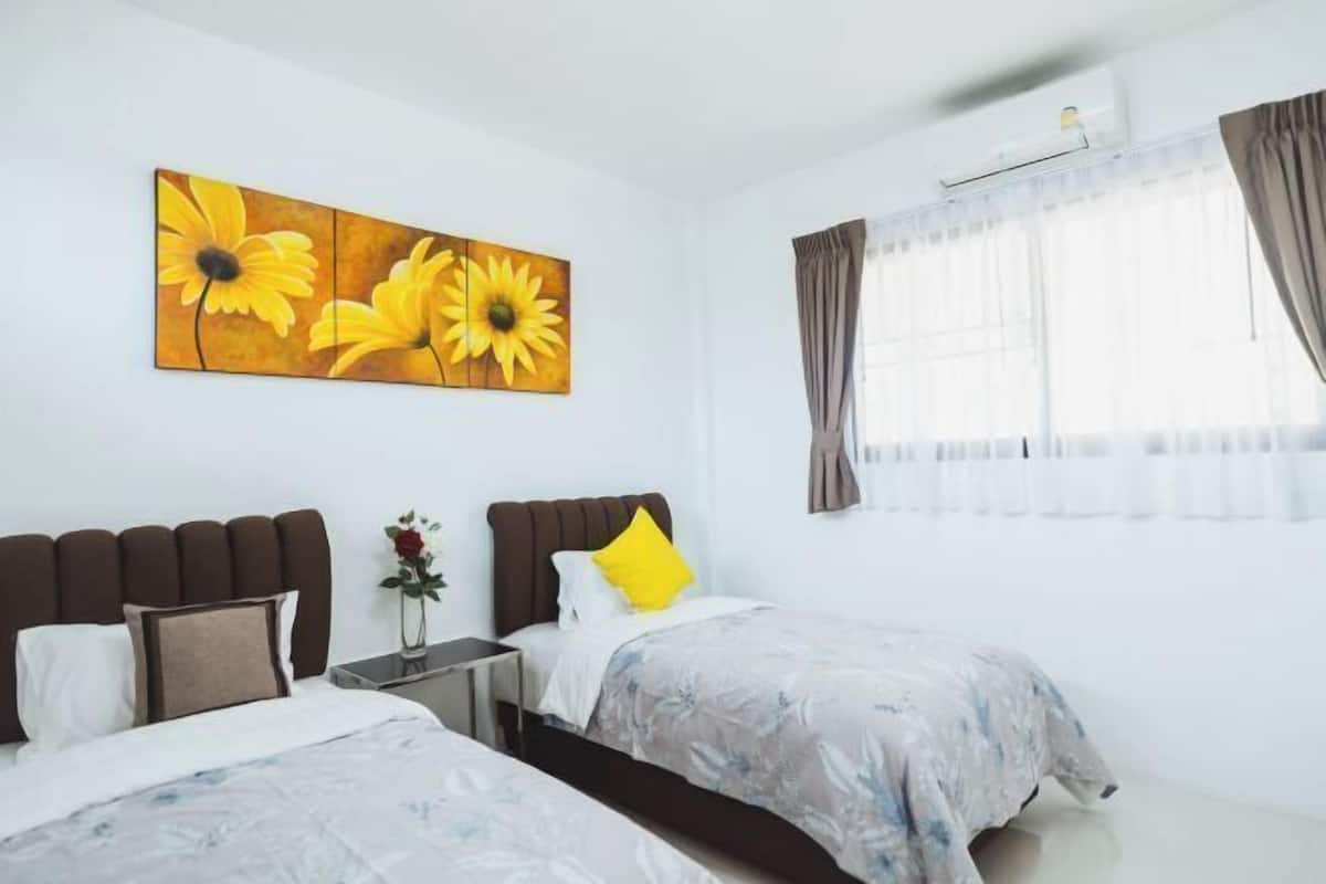 Una camera d'albergo con due letti, un quadro grande di girasoli sulla parete, una finestra con tende e un tavolino con un vaso di fiori.