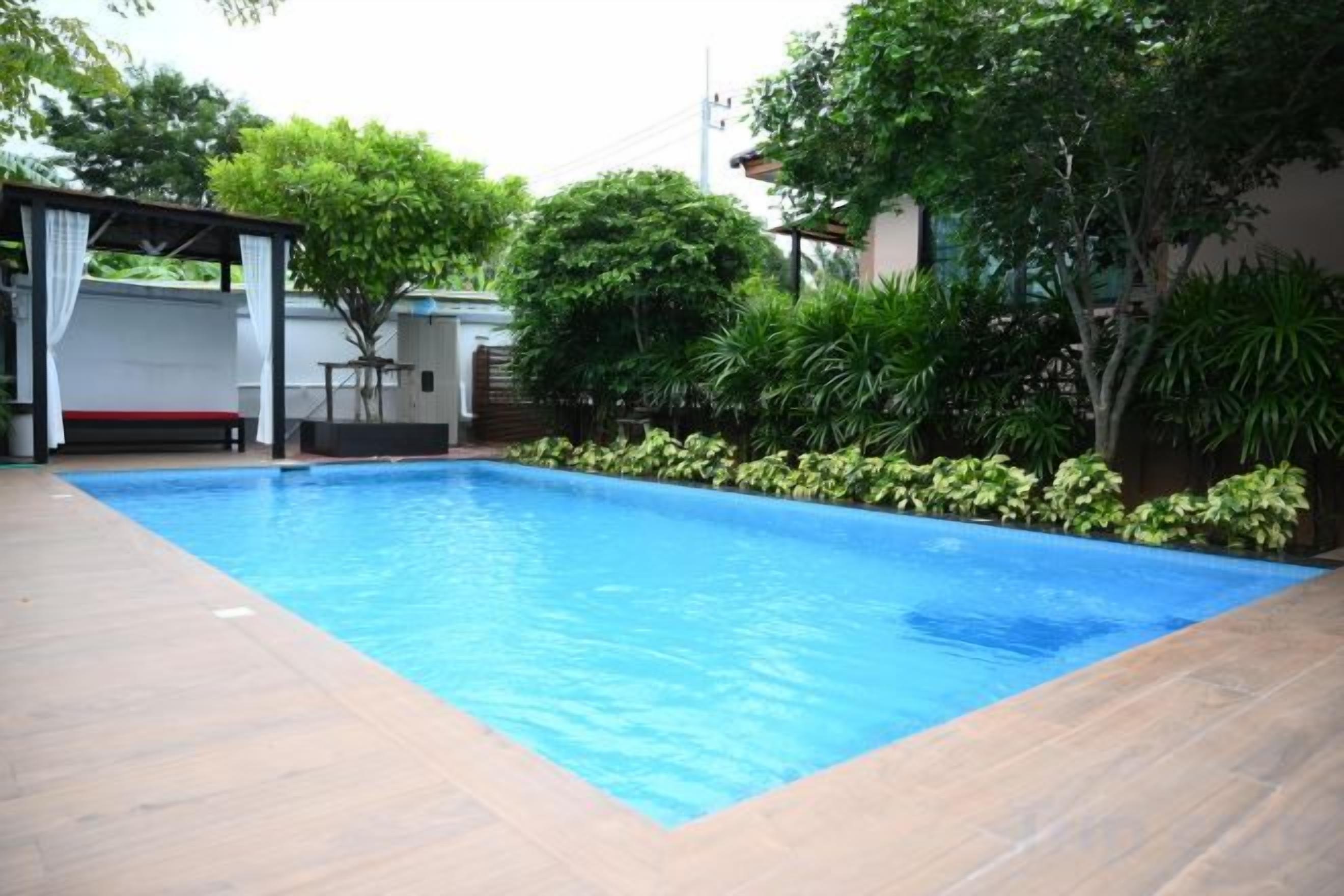 A rectangular swimming pool with a tiled deck, surrounded by greenery and a covered seating area.