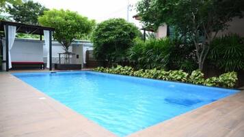 A rectangular swimming pool with a tiled deck, surrounded by greenery and a covered seating area.