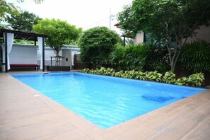 Una piscina rectangular con un borde de baldosas, rodeada de vegetación y una zona de estar cubierta.