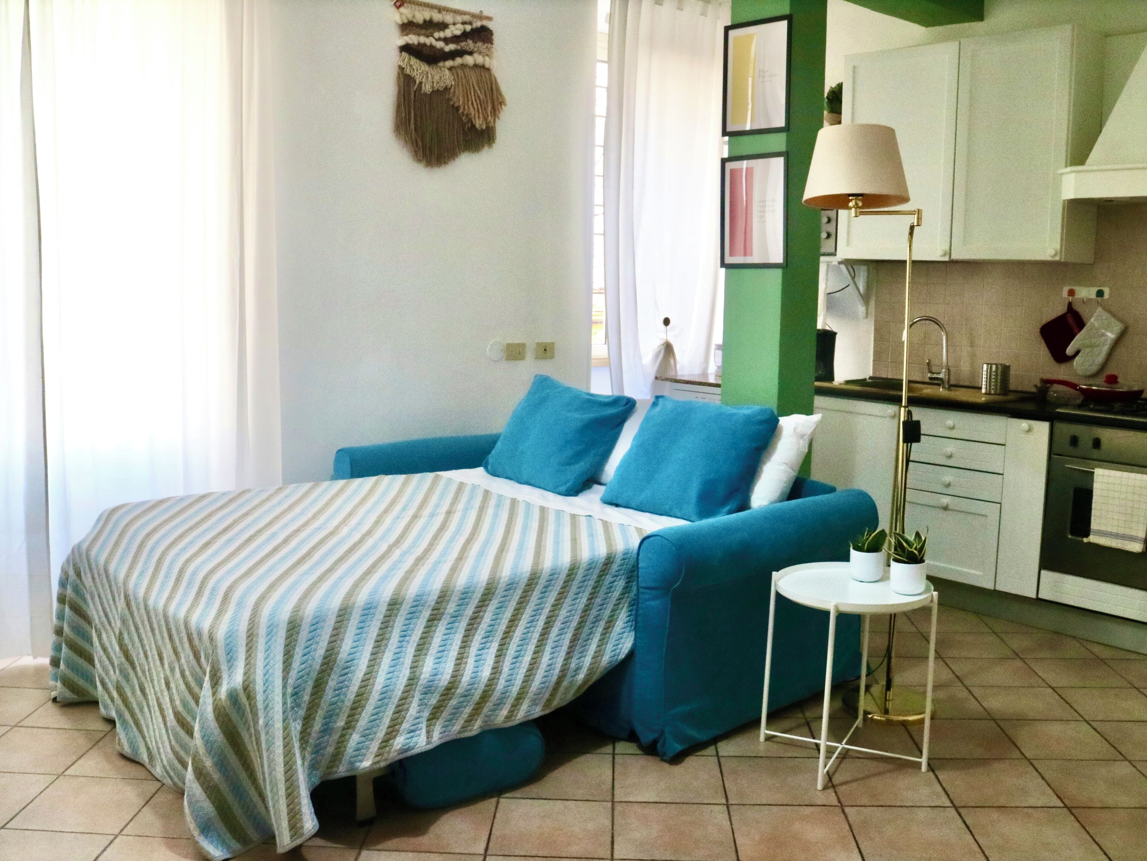 A bedroom with a bed, a bedside table, a lamp, and a kitchenette in the background.