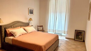 Ein Schlafzimmer mit Bett, Sessel, Fenster mit Vorhängen, Lampe und einem gerahmten Bild an der Wand.