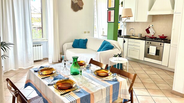 Eine helle, moderne Küche mit weißen Schränken, einem Esstisch für vier Personen und einer gemütlichen Wohnbereits mit Sofa und blauen Kissen.