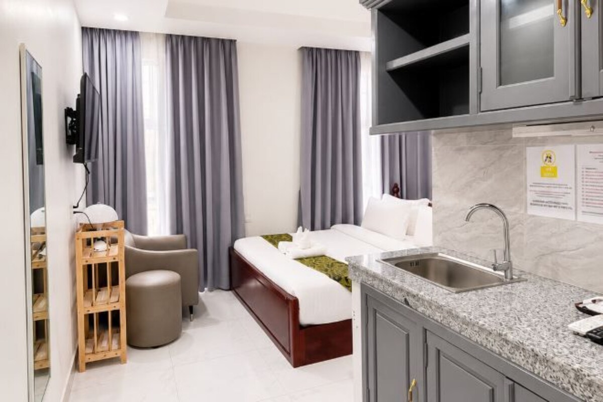 A hotel room with a bed, a desk, a chair, a small table, a sofa, a TV, a kitchen area with a sink, and a cabinet.