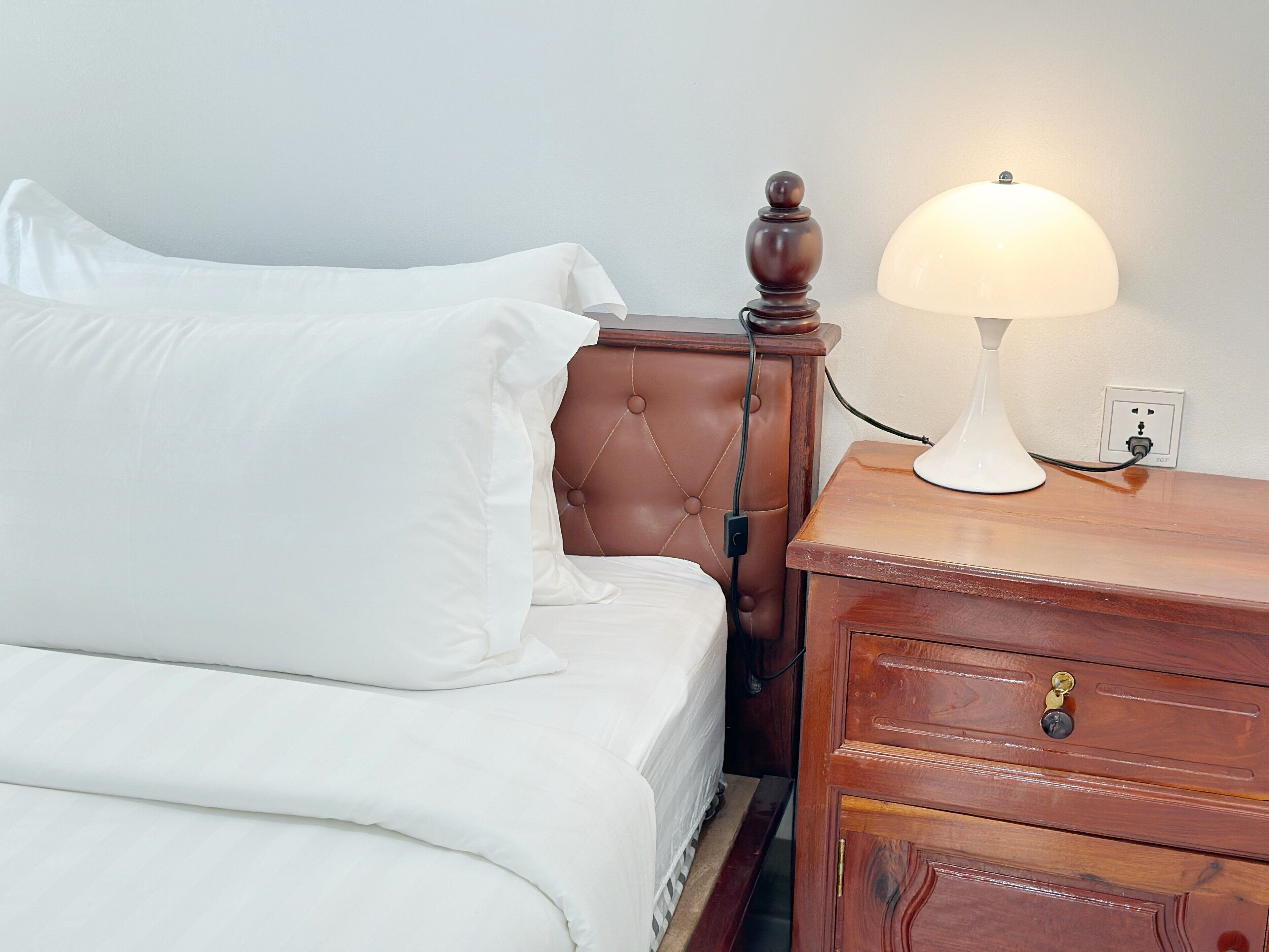 A neatly made bed with white linens, a wooden nightstand with a lamp, and a wall socket.