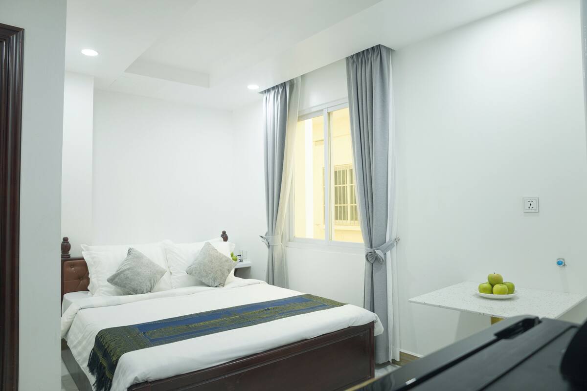 Een slaapkamer met een bed, nachtkastje, een klein tafeltje met appels en een raam met gordijnen.