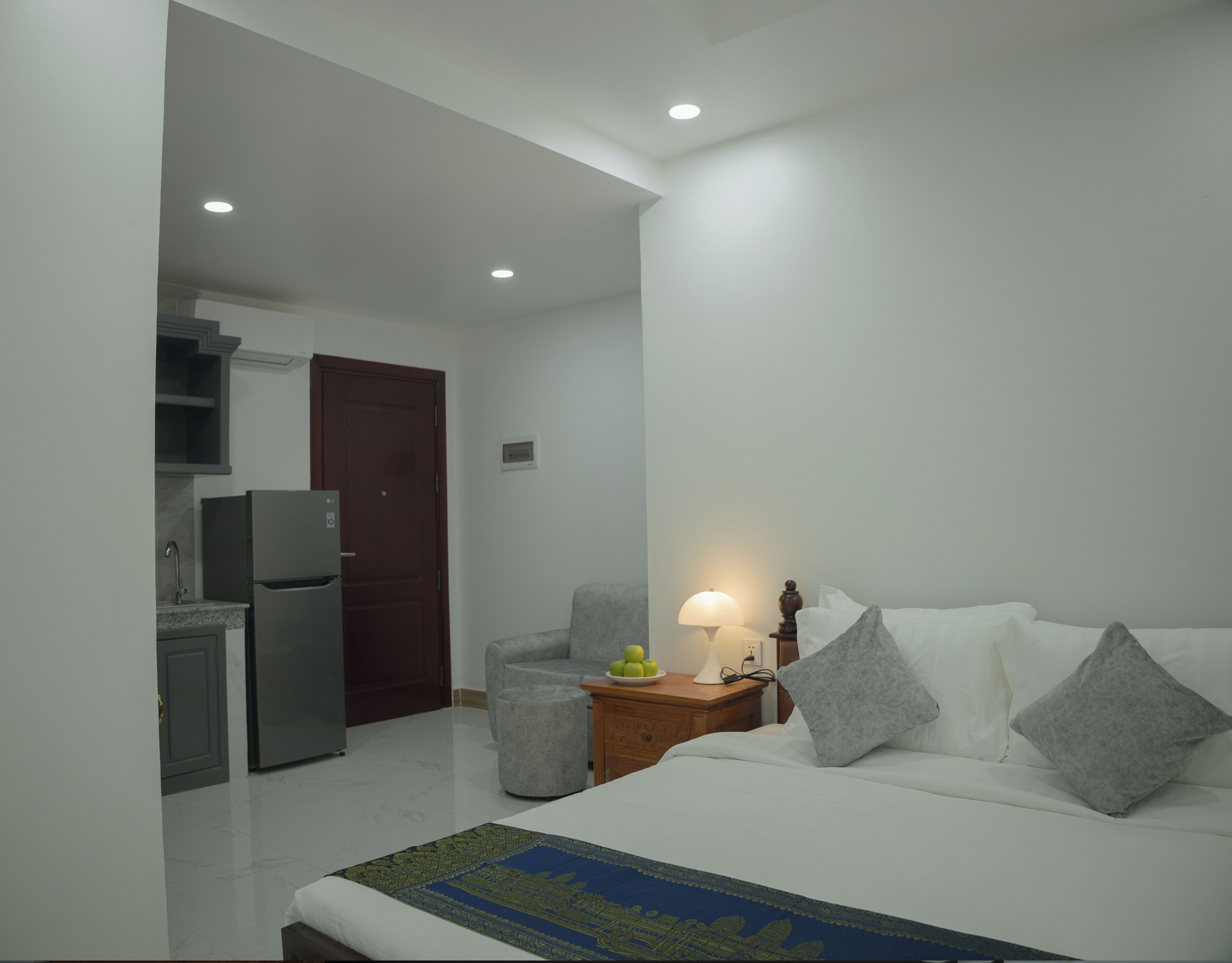 Habitación de hotel con cama, mesita de noche con lámpara, sofá, pequeña mesa con frutas, refrigerador y área de cocina con fregadero y armarios.