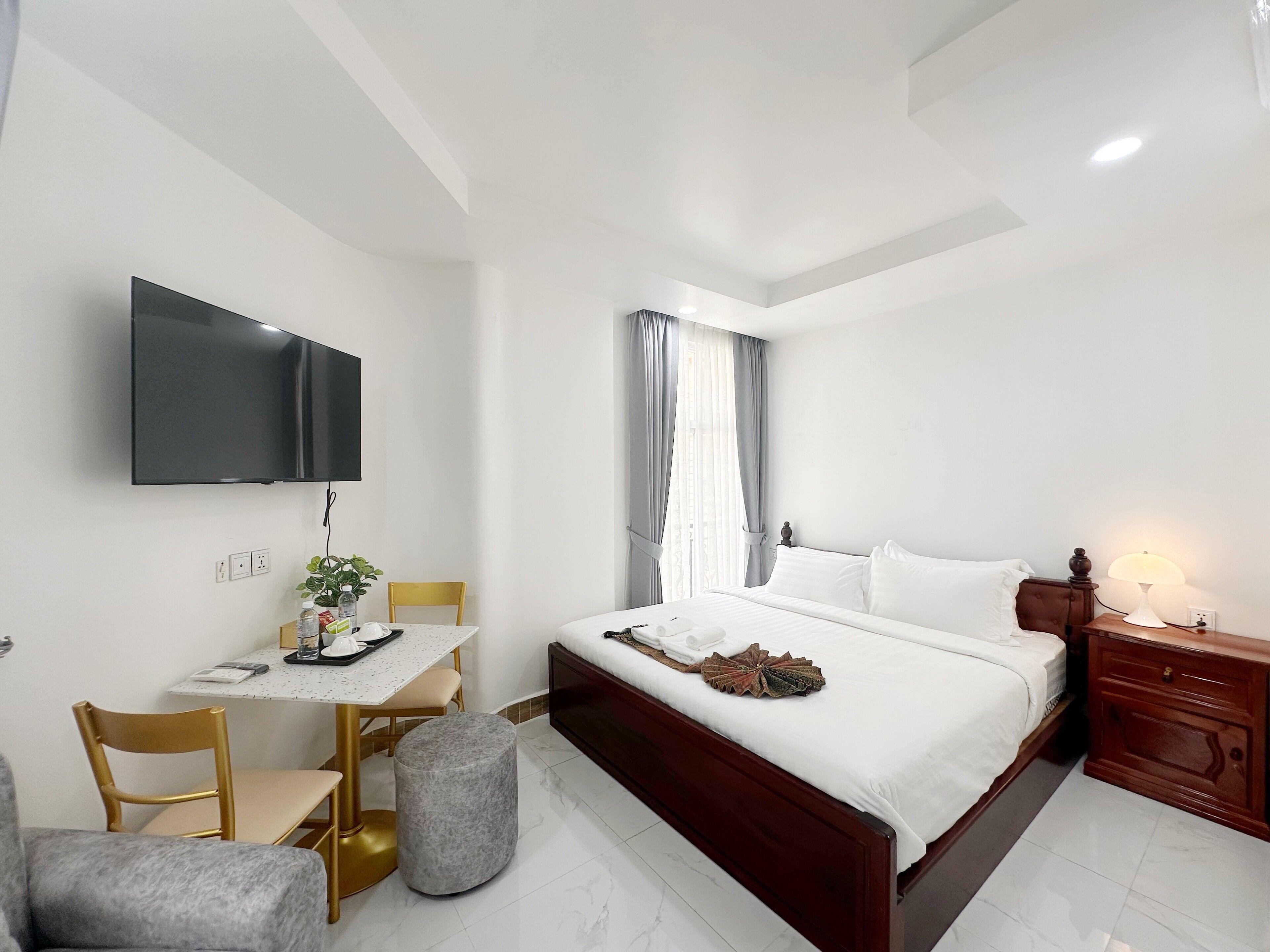 A hotel room with a bed, a desk with a chair, a television mounted on the wall, and a nightstand with a lamp.