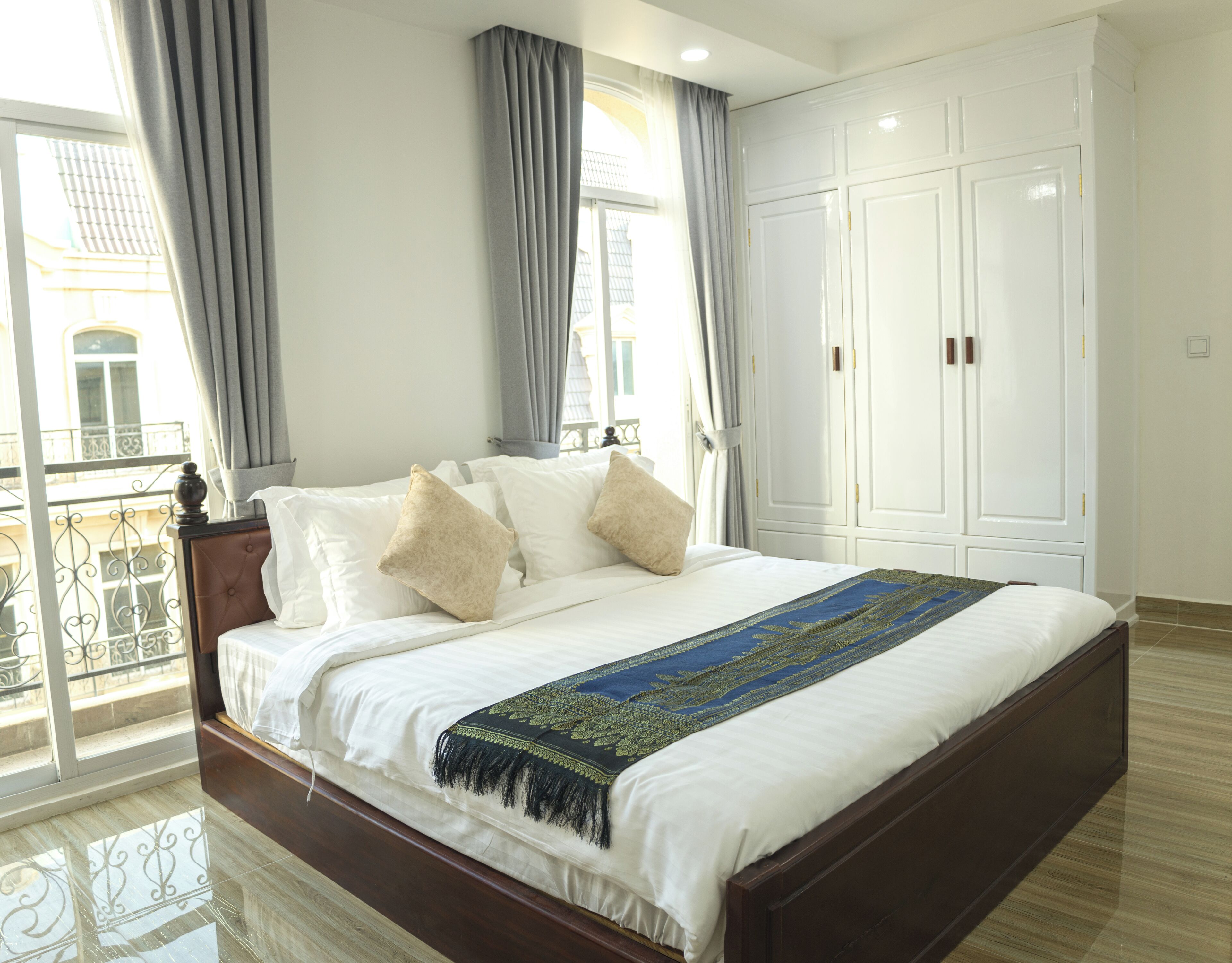 A neatly made bed with a white comforter and decorative pillows, a wooden headboard, and a large window with curtains.