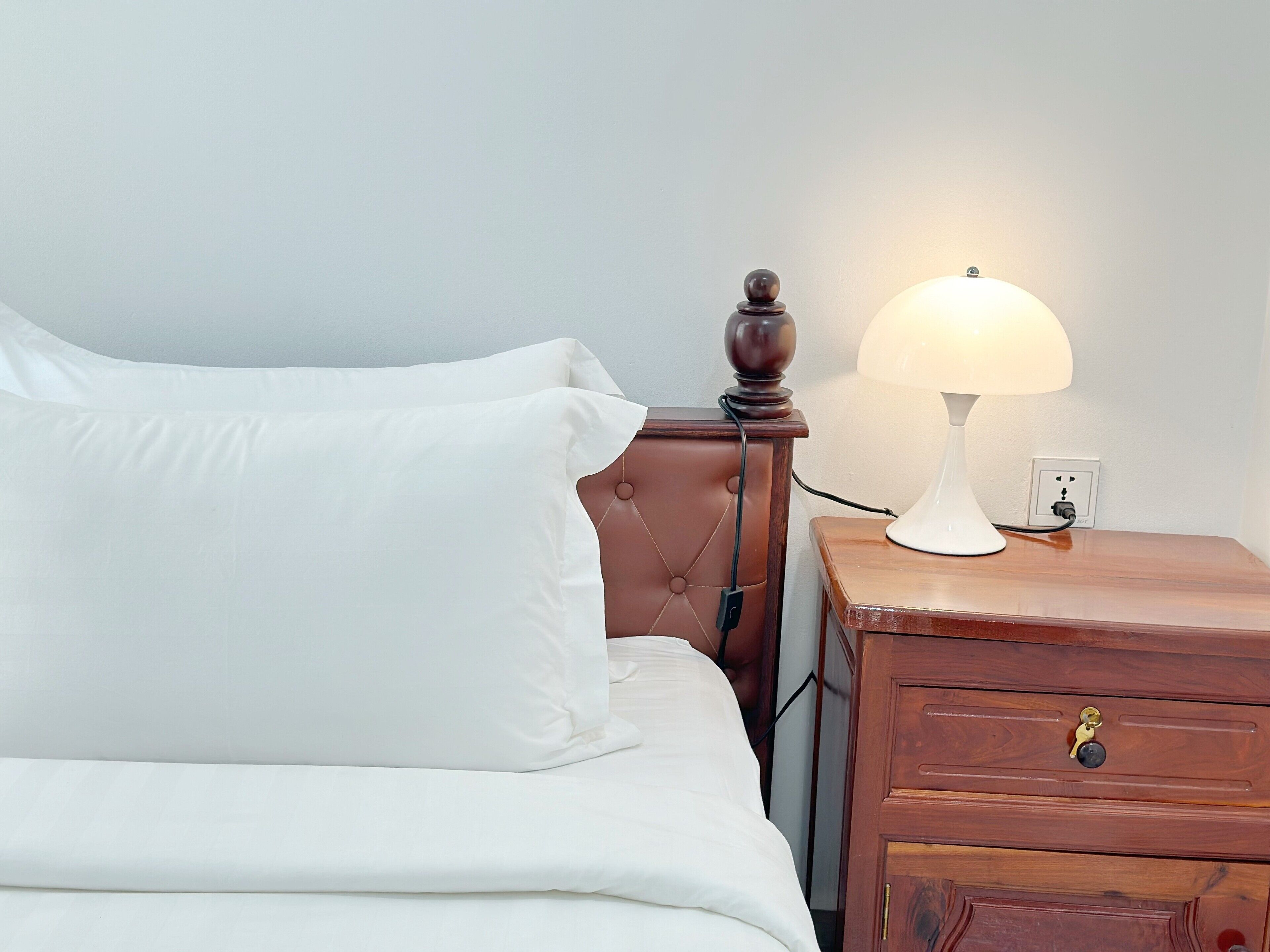 A neatly made bed with white linens, a wooden nightstand with a lamp, and a drawer with a brass handle.
