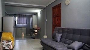 Un salon avec un canapé, un fauteuil, un coin cuisine à l’arrière-plan et une porte en bois.