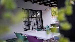 Un espace extérieur aménagé avec des tables blanches et des chaises vertes, une grande fenêtre aux montures noires et un mur en briques blanches.