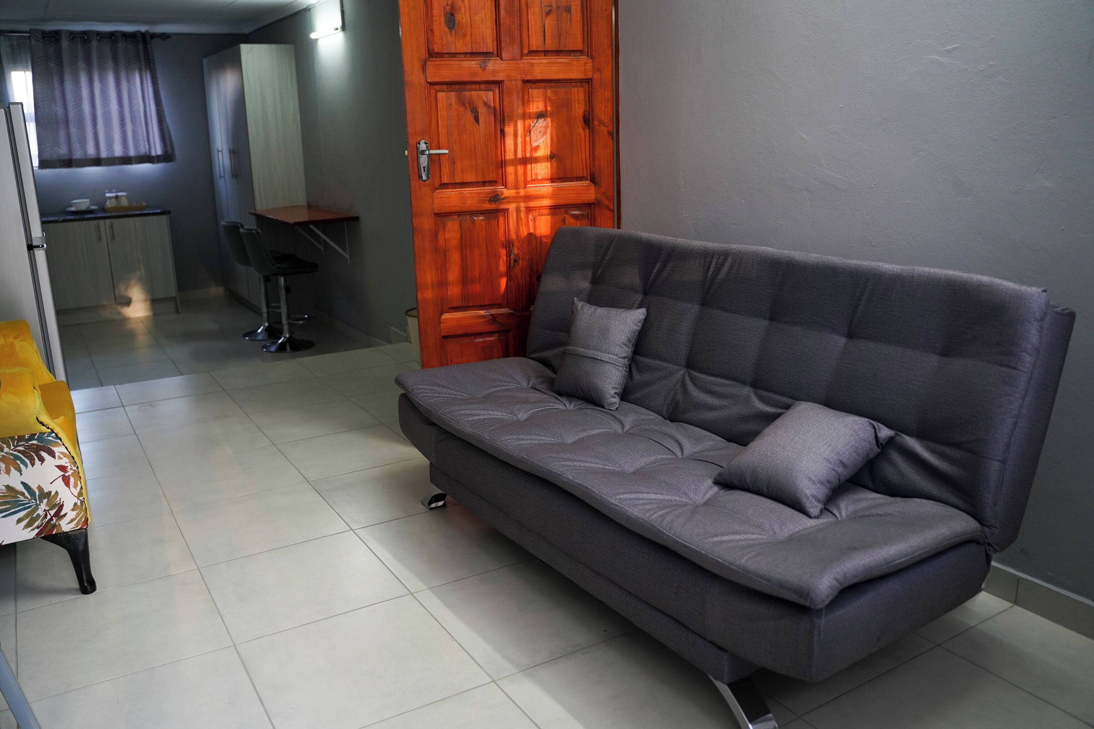 A grey sofa with a cushion, a wooden door, and a kitchen area with cabinets and a countertop.