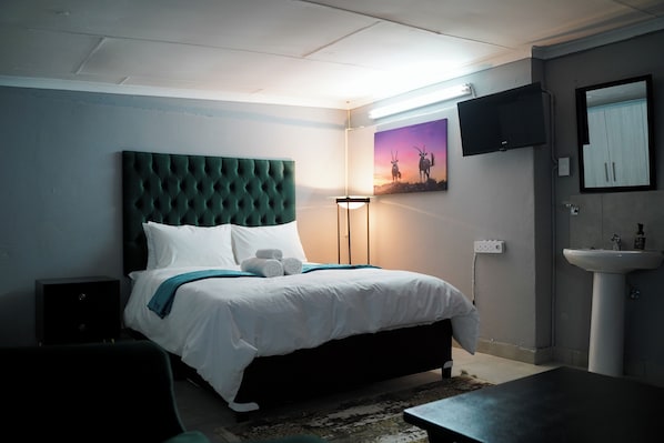 A bedroom with a large bed, a bedside table, a floor lamp, a painting on the wall, a TV mounted above the bed, and a sink in the corner. - Aqua Guest House (Sandton)