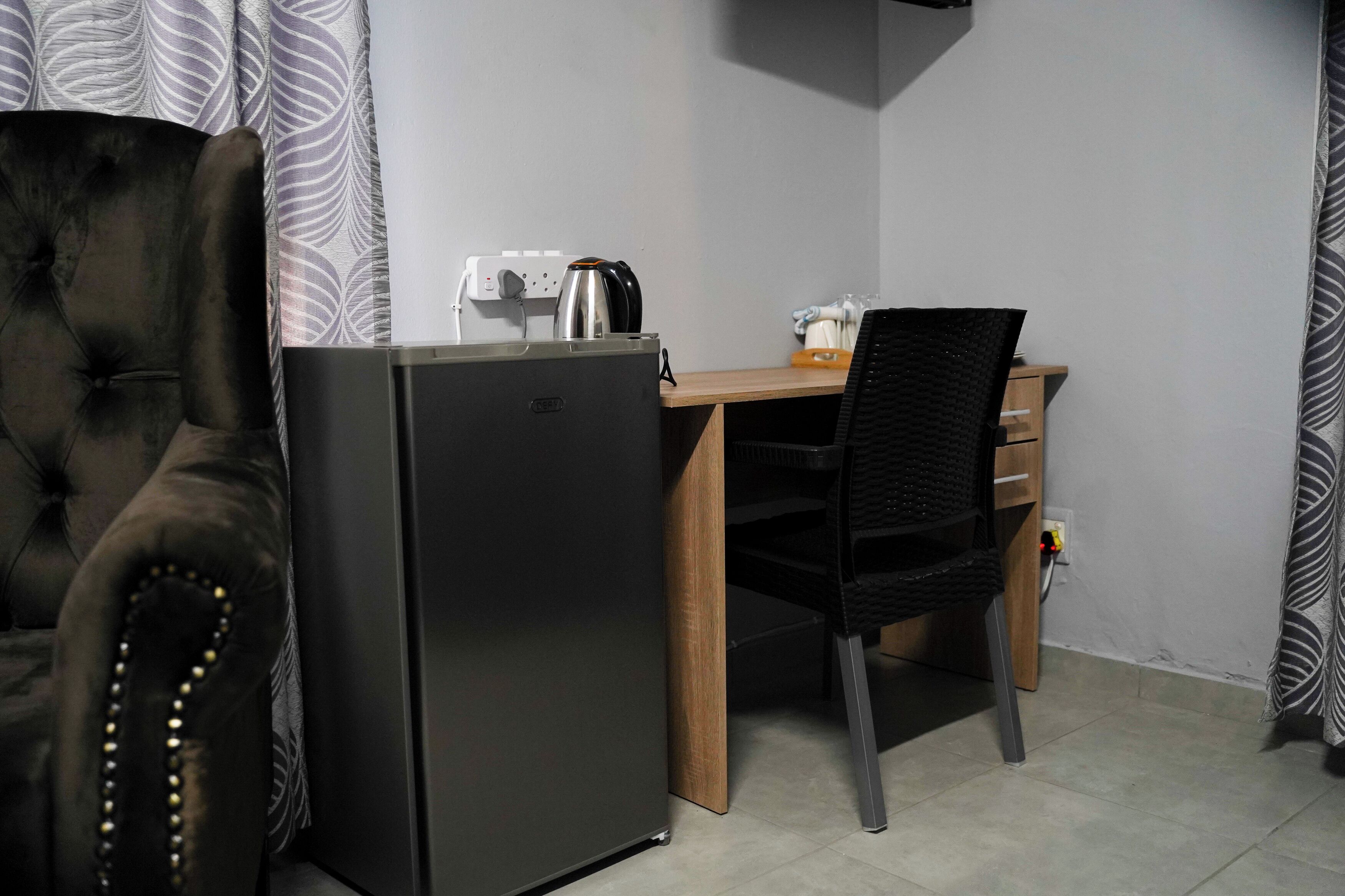 Habitación compacta con sofá oscuro, escritorio pequeño de madera, silla negra y refrigerador negro.