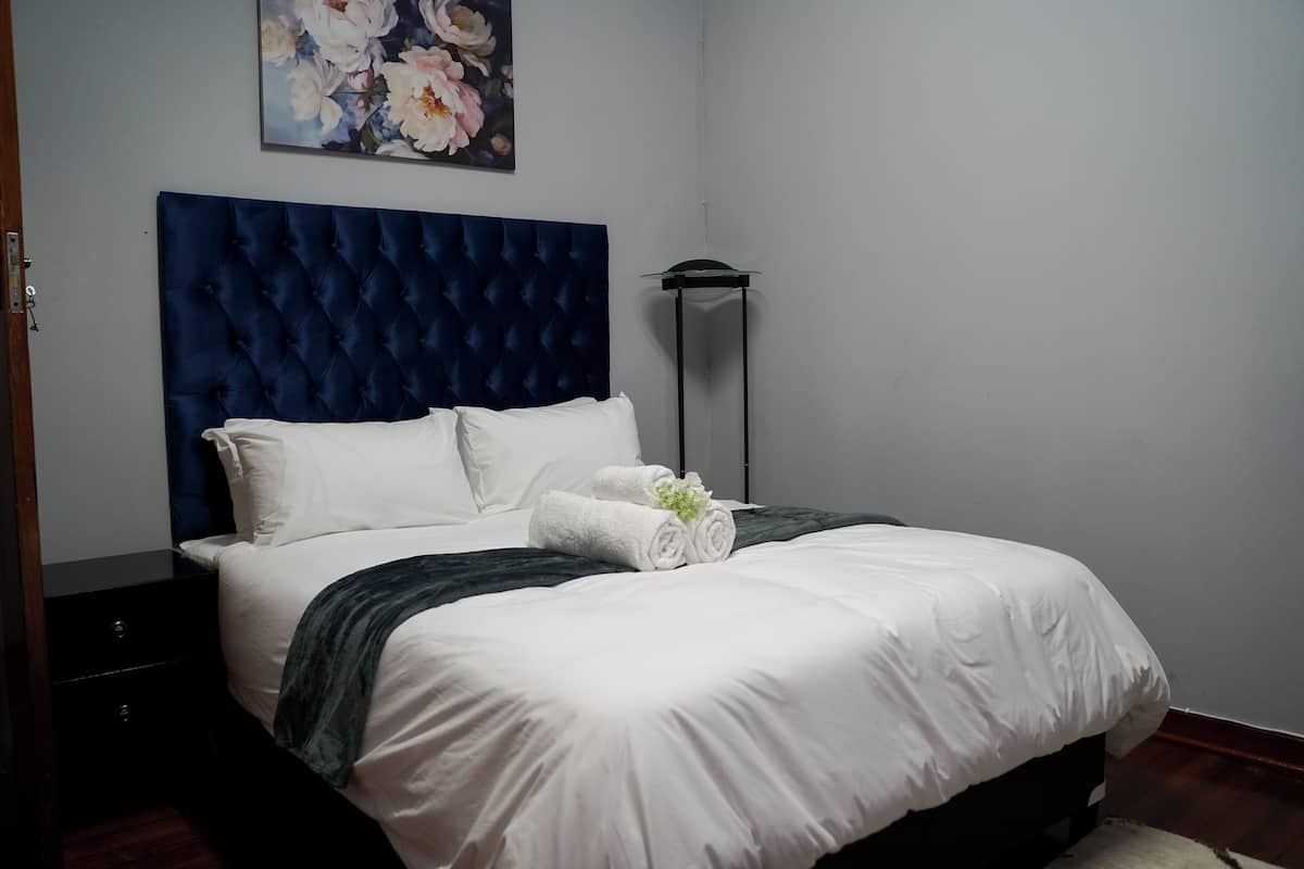 Ein ordentlich bezogenes Bett mit weißen Leinen und einem dunkelblauen Kopfteil, eine Stehlampe und ein Blumenbild an der Wand.