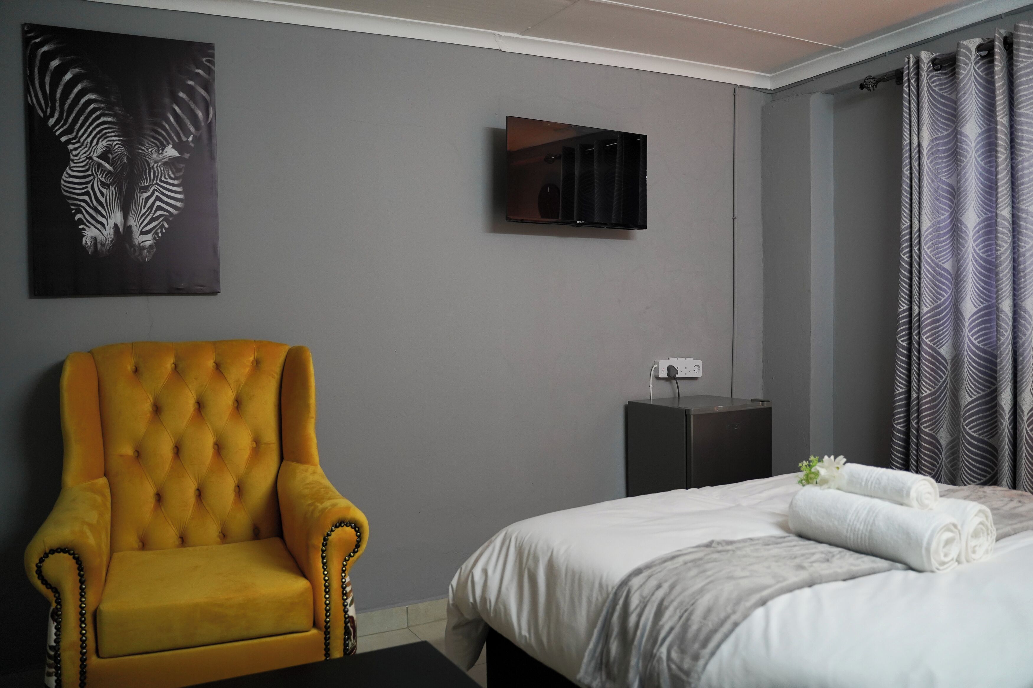 Een hotelkamer met een gestoffeerde gele fauteuil, een bed met witte lakens, een wandtelevisie en een kamerbreed gordijn met dessin.