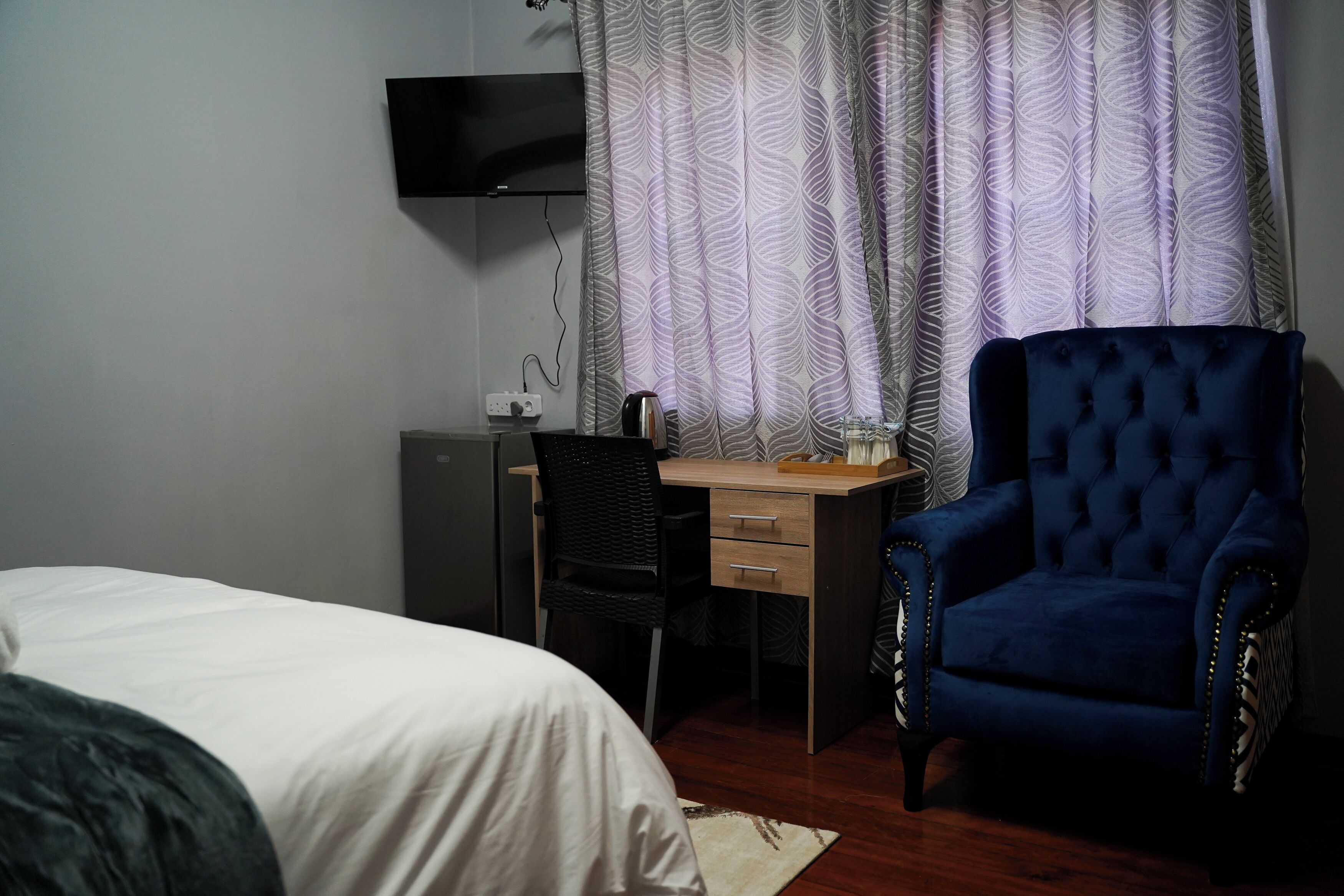 A bedroom with a bed, a desk with a chair, a blue armchair, a television mounted on the wall, and patterned curtains.