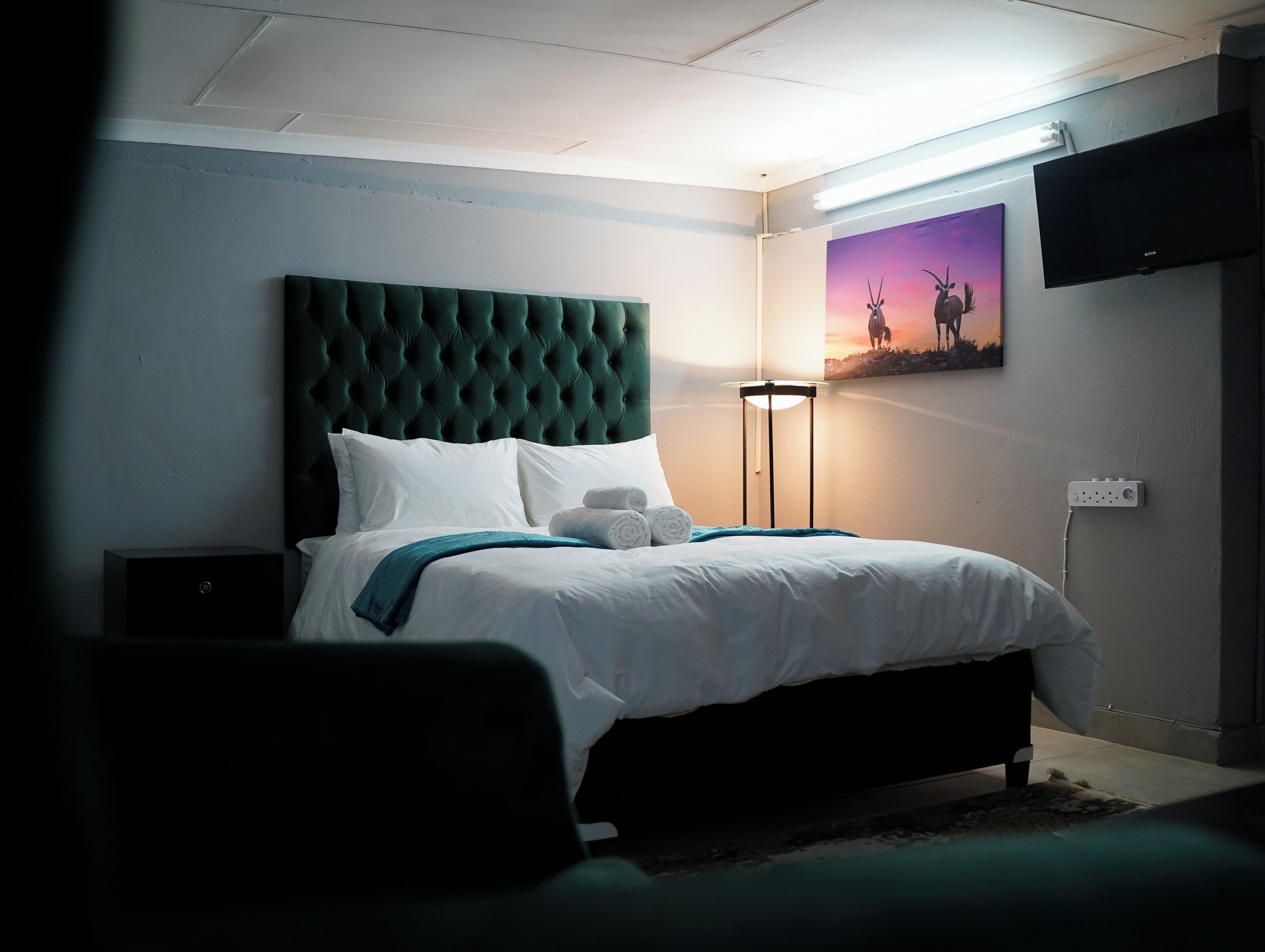 A neatly made bed with white linens and a green headboard, a bedside lamp, and a painting of horses on the wall.
