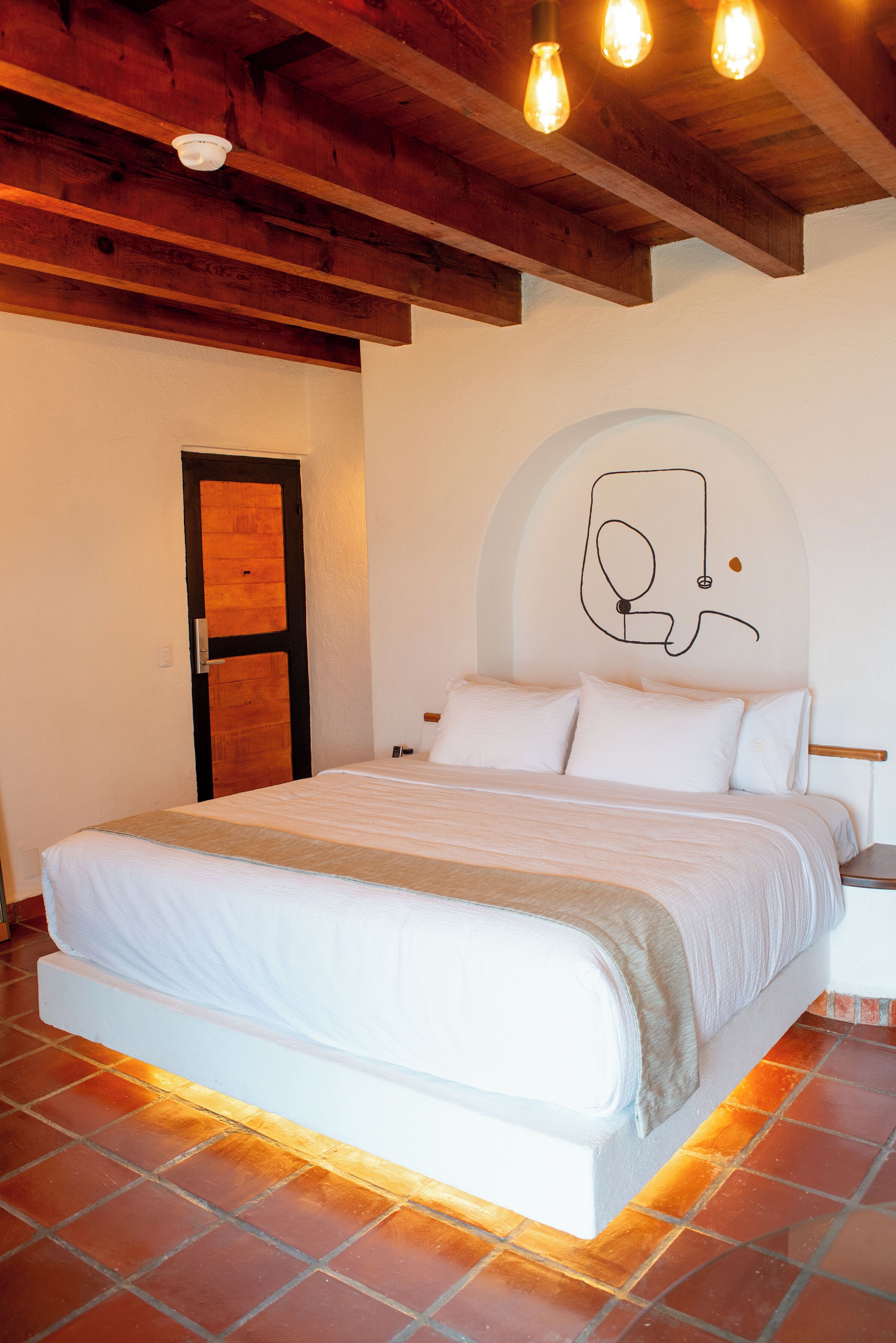 Un dormitorio con cama, vigas de madera en el techo, una puerta blanca y un muro con un dibujo lineal sencillo.