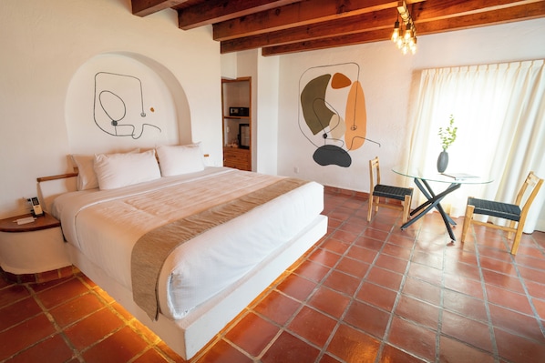 A bedroom with a large bed, a wooden headboard, a nightstand, a bedside table, a glass table with chairs, and a window with curtains. - Hanna Hotel Boutique (Valle de Bravo)