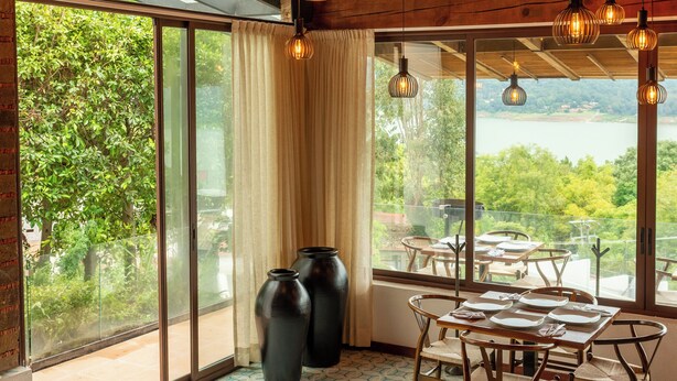 A spacious room with a large dining table, wooden beams, and large windows offering a view of greenery and a body of water.