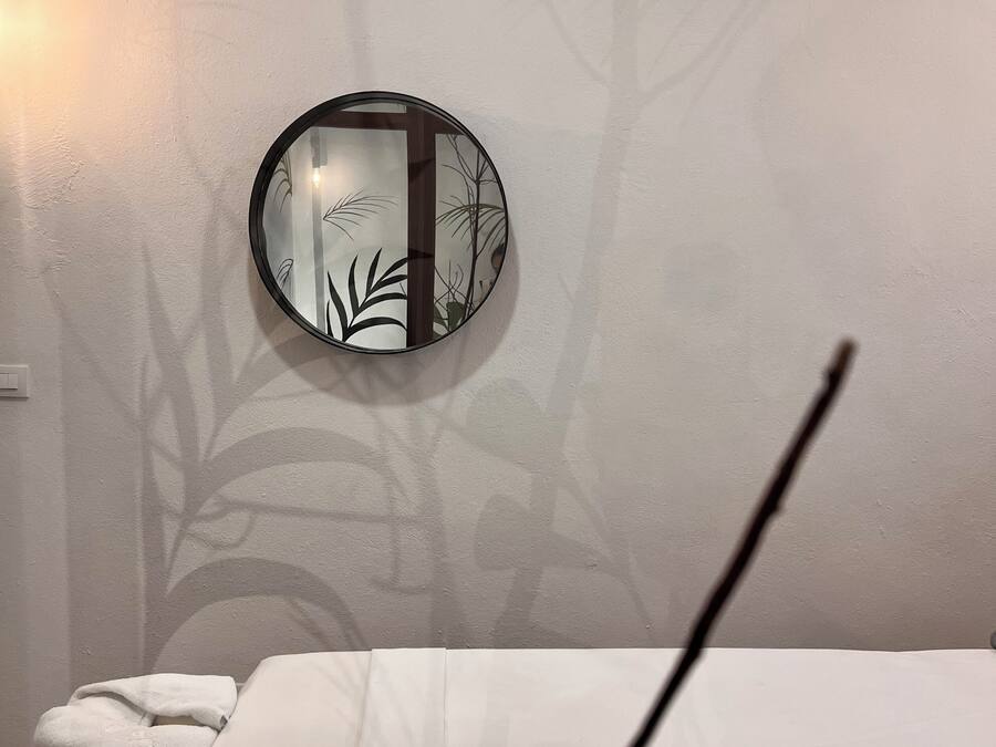 Una habitación con un espejo redondo en la pared, una cama con colcha blanca y una vista parcial de un baño con toalla y una silla blanca.