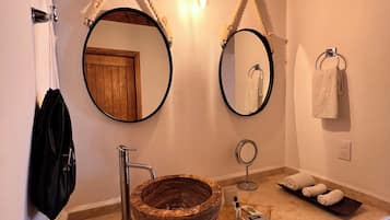 A bathroom with a wooden vanity, a round marble sink, two wall-mounted mirrors, a towel rack, and a small shelf with toiletries.