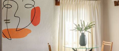 A bedroom with a bed, a glass table with a vase, a chair, and a wall with abstract art.