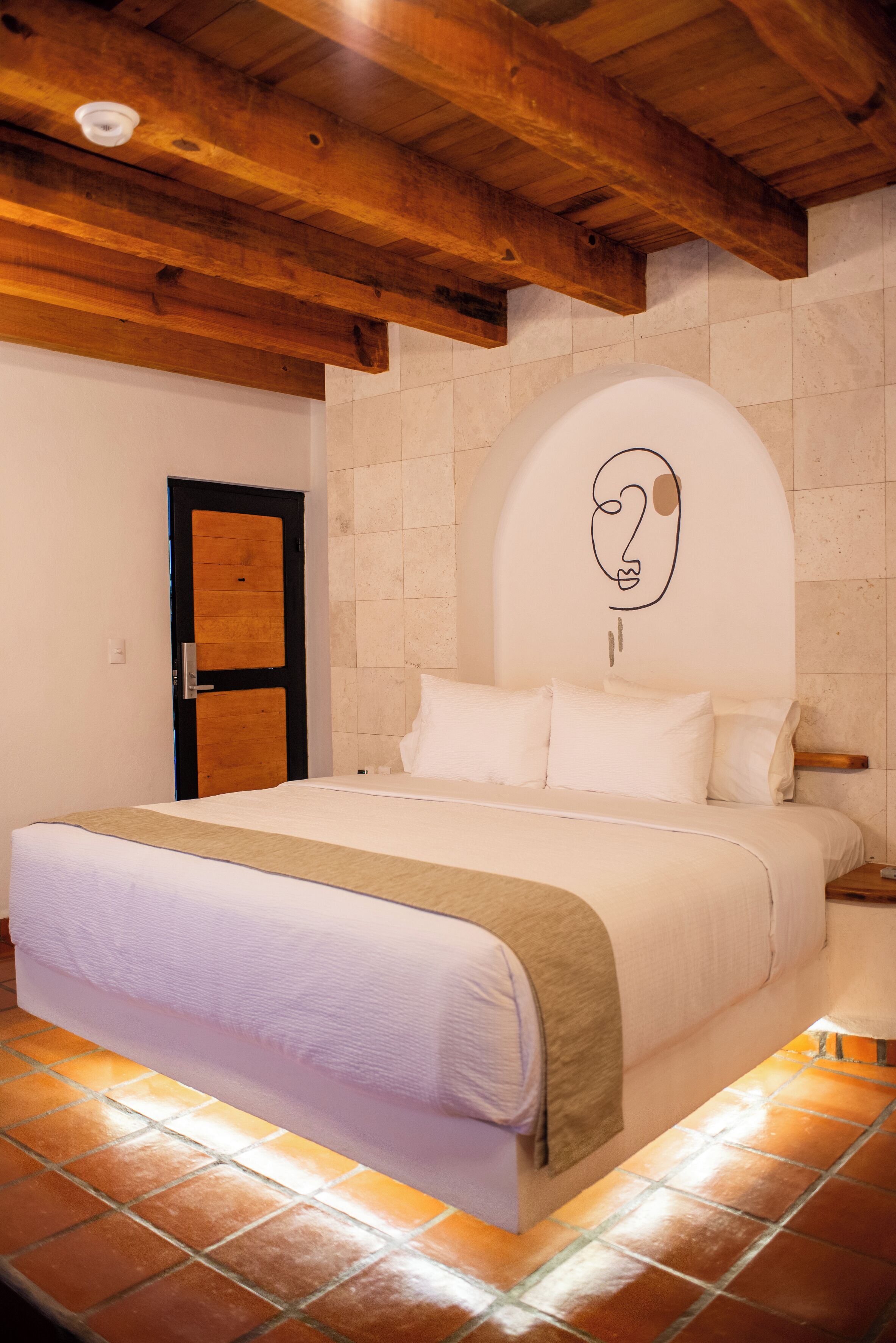 A bedroom with a large bed, wooden ceiling beams, a tiled floor, and a simple wall design.
