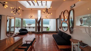 A spacious room with a black sofa, wooden chairs, a wooden table, and a large vase. There are multiple mirrors on the walls and a ceiling light fixture.