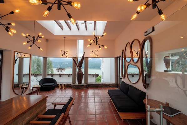 Una habitación amplia con un sofá negro, sillas de madera, una mesa de madera y un jarrón grande. Hay varios espejos en las paredes y una lámpara en el techo.