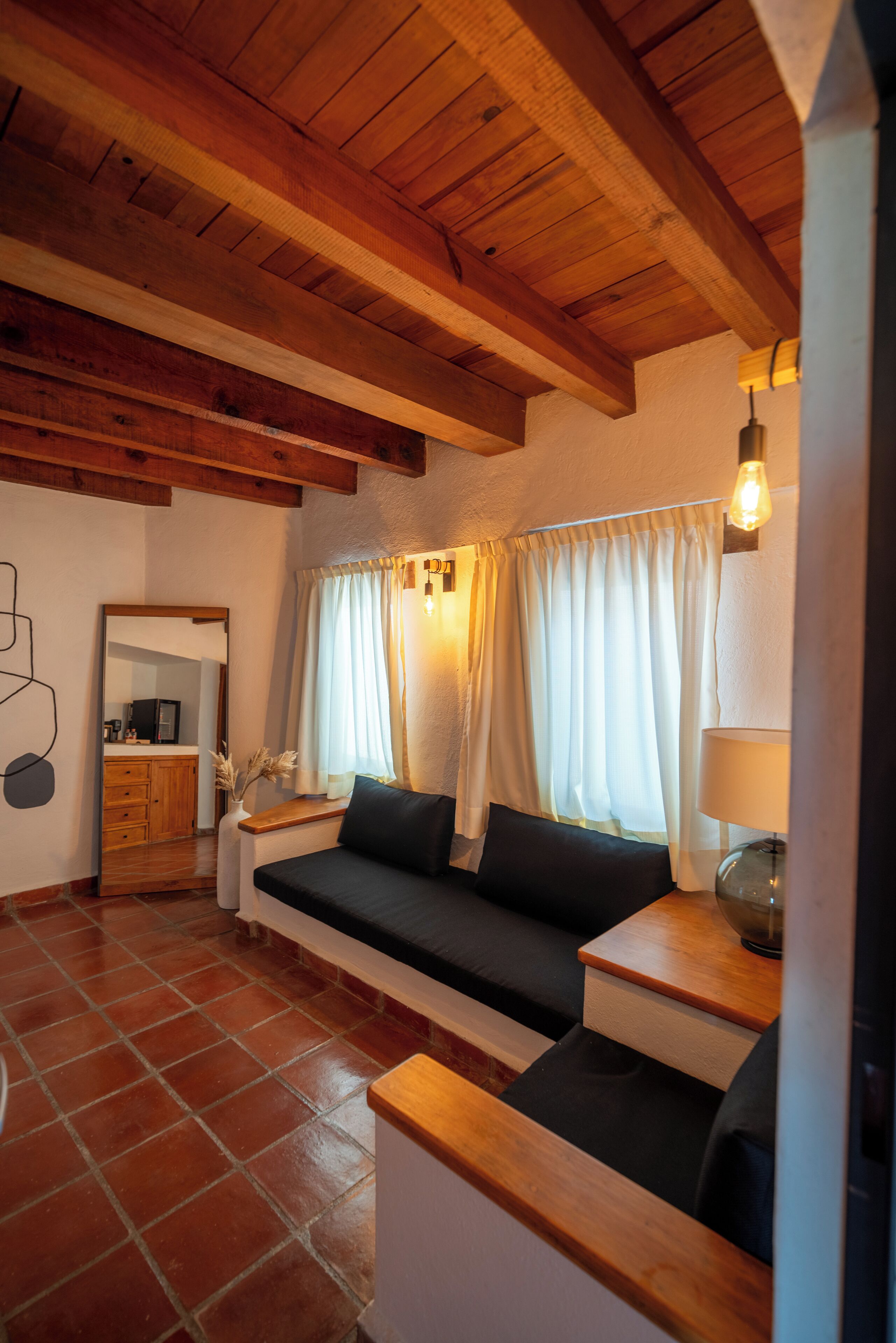 A cozy living room with a wooden ceiling, a sofa, a small table with a lamp, and a window with curtains.