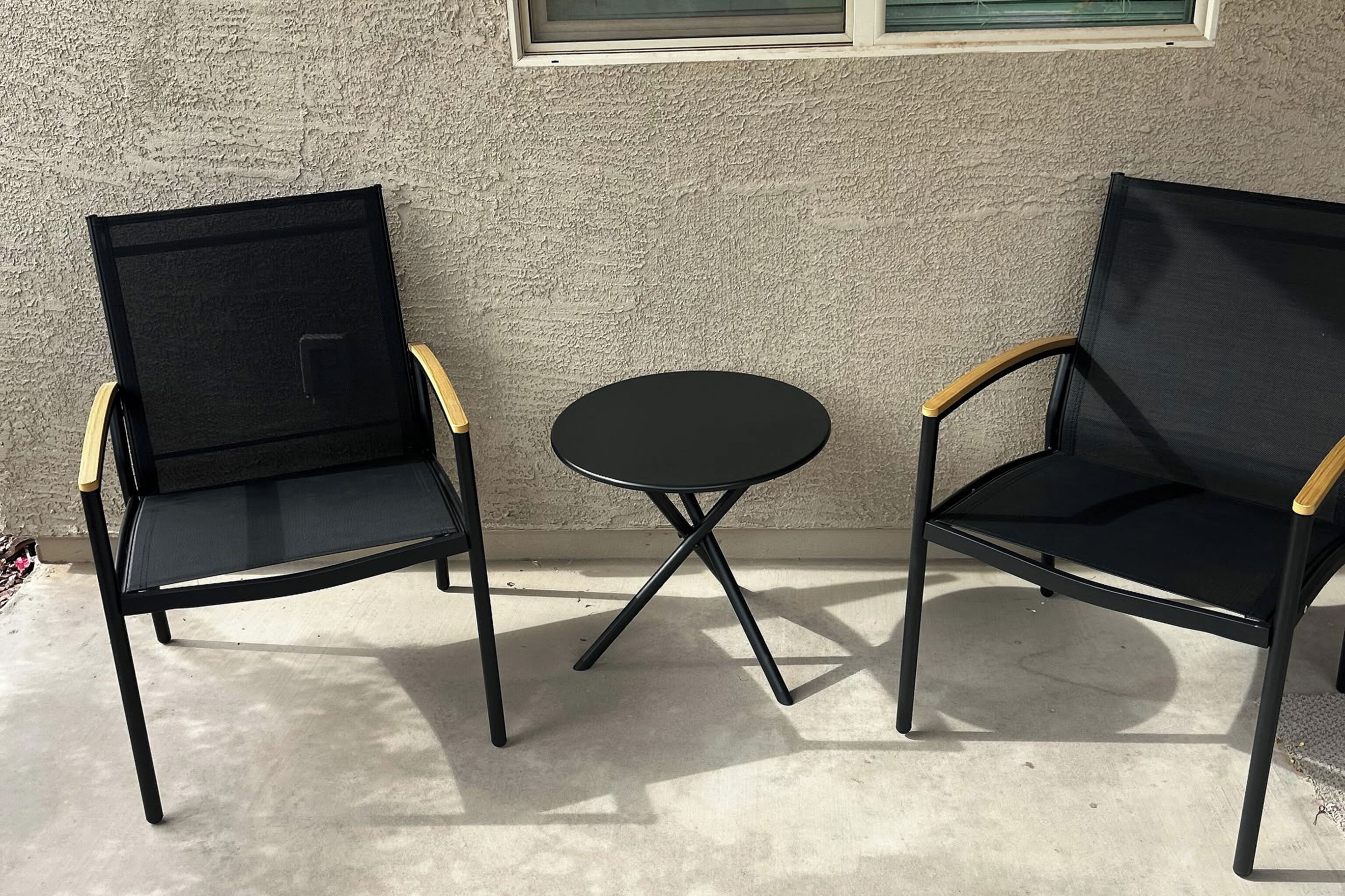 Dos sillas negras con reposabrazos de madera y una pequeña mesa redonda entre ellas, ubicadas frente a una pared con textura y una ventana en la parte superior.