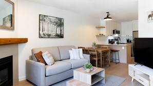 A modern living room with a grey sofa, a coffee table, and a TV stand. There is a kitchen area with a dining table and chairs.