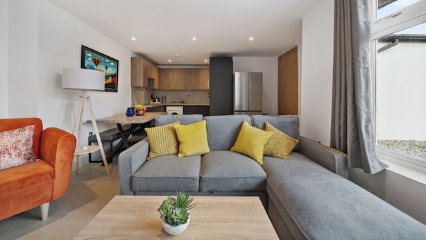 Een moderne woonkamer met een grijze modulaire bank, een kleine oranje bank en een houten salontafel met een plant. In de achtergrond is een keuken zichtbaar met houten kastjes en een raam met grijze gordijnen.