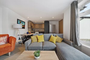 Ein modernes Wohnzimmer mit einem grauen Ecksofa, einem kleinen orangefarbenen Sofa und einem Couchtisch aus Holz mit einer Pflanze. Im Hintergrund ist eine Küche mit Holzschränken und einem Fenster mit grauen Vorhängen erkennbar.