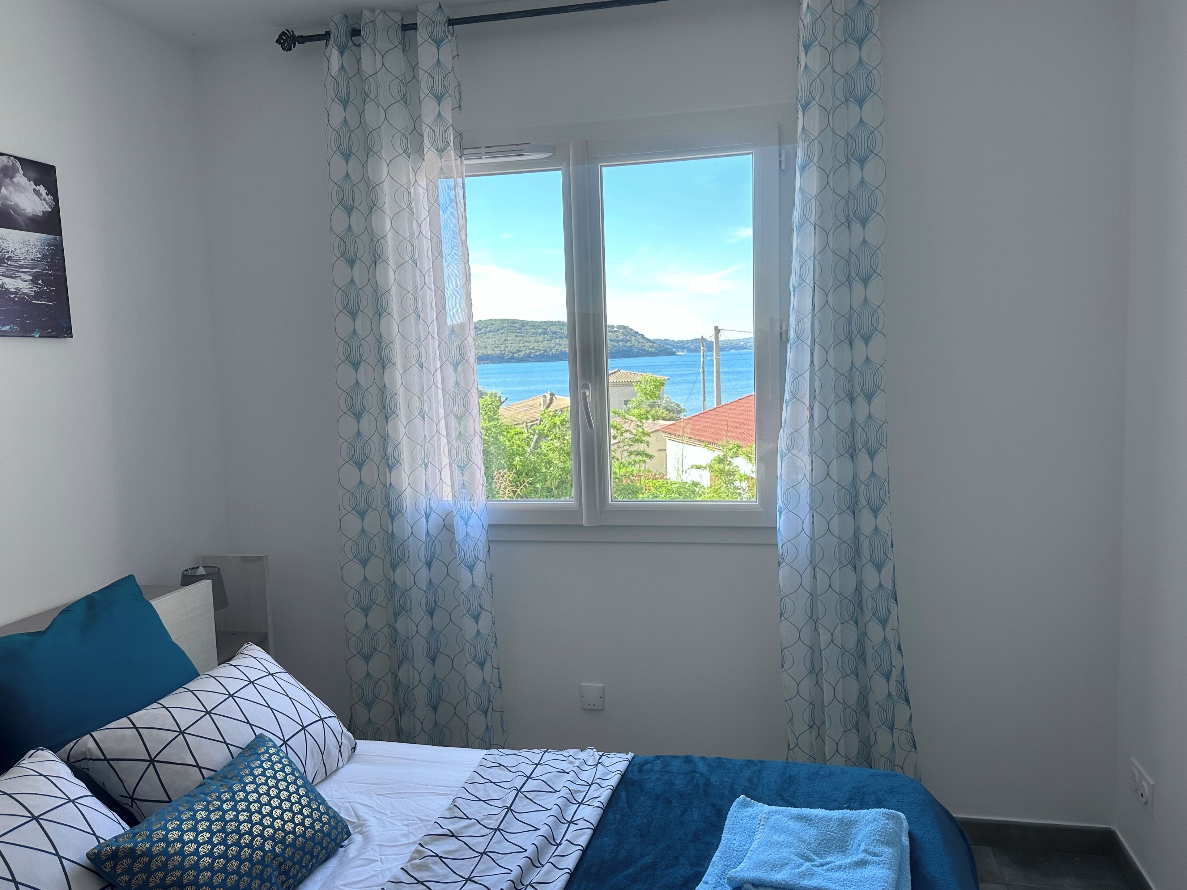 Un dormitorio con cama, ventana con cortinas, vista al mar y un cuadro en la pared.