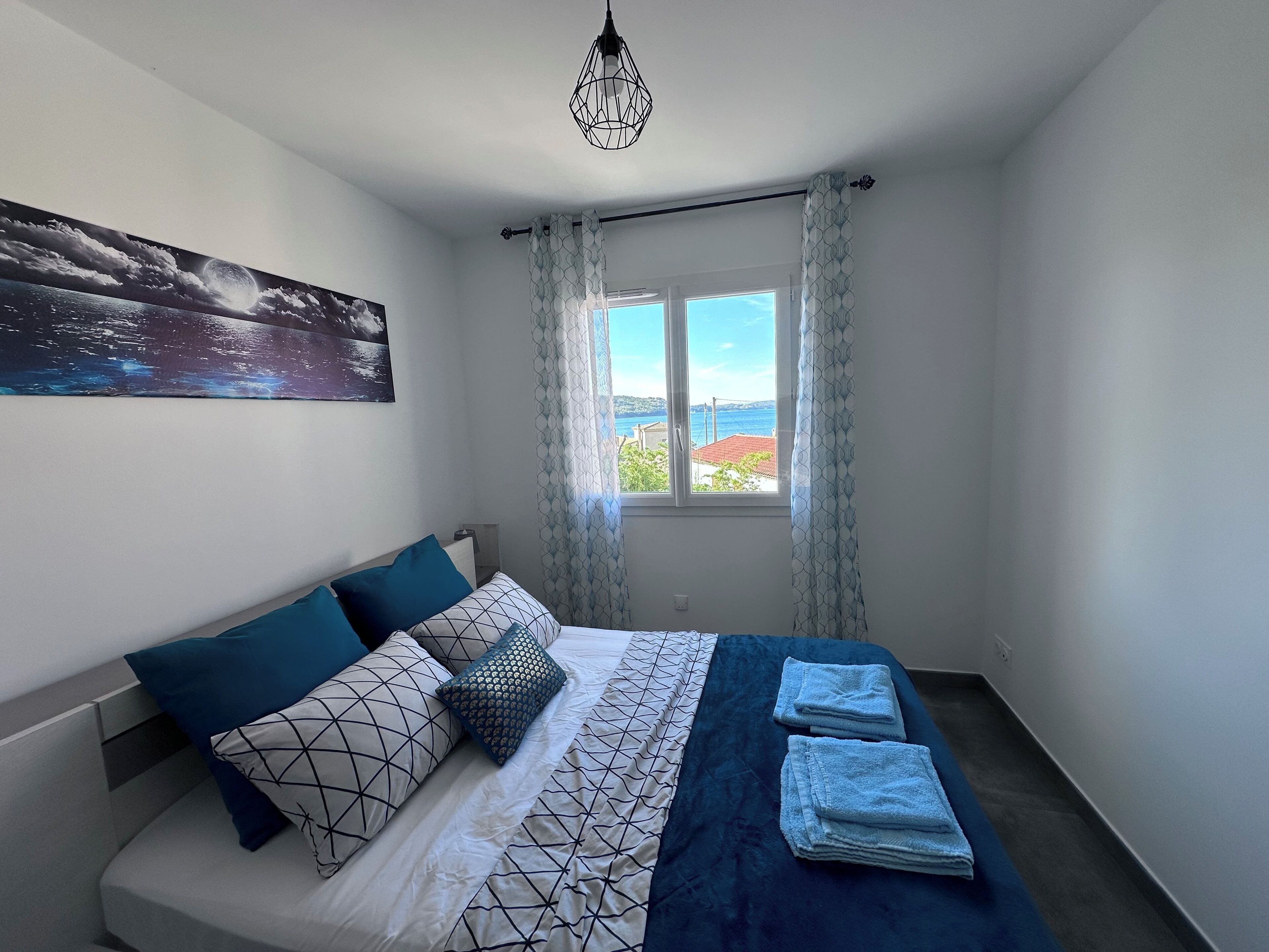 A bedroom with a bed, a painting, a window with curtains, and a view of the sea.