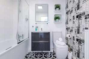 A bathroom with a bathtub, a glass shower enclosure, a white toilet, and a sink with a mirror above it. There are two small shelves with plants on the wall. - NEW! Stunning City Centre Flat, Amazing Location! (Chester)