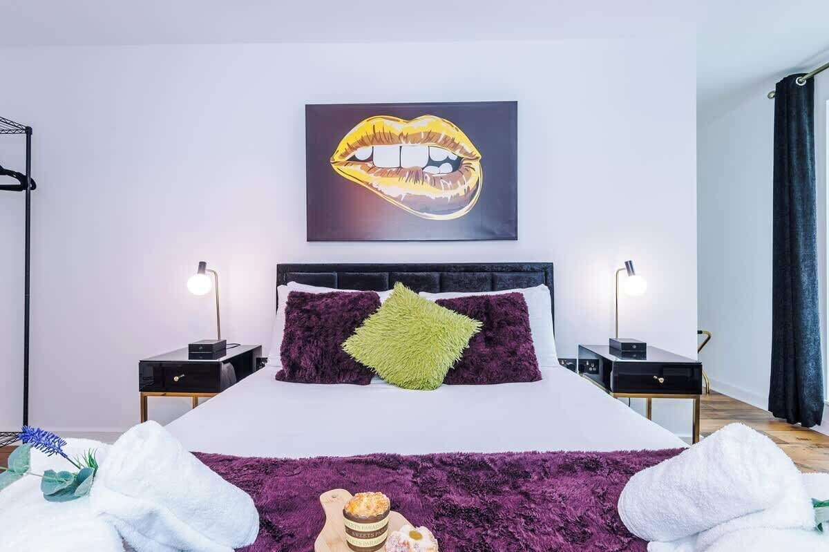 A modern bedroom with a large bed, two bedside tables, a wall-mounted artwork of lips, and a purple throw blanket.