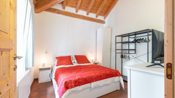 A bedroom with a wooden ceiling, a bed with red bedding, a white bedside table, a nightstand, a wardrobe, and a desk with a chair. - Campari Aperol in Iseo (Iseo)
