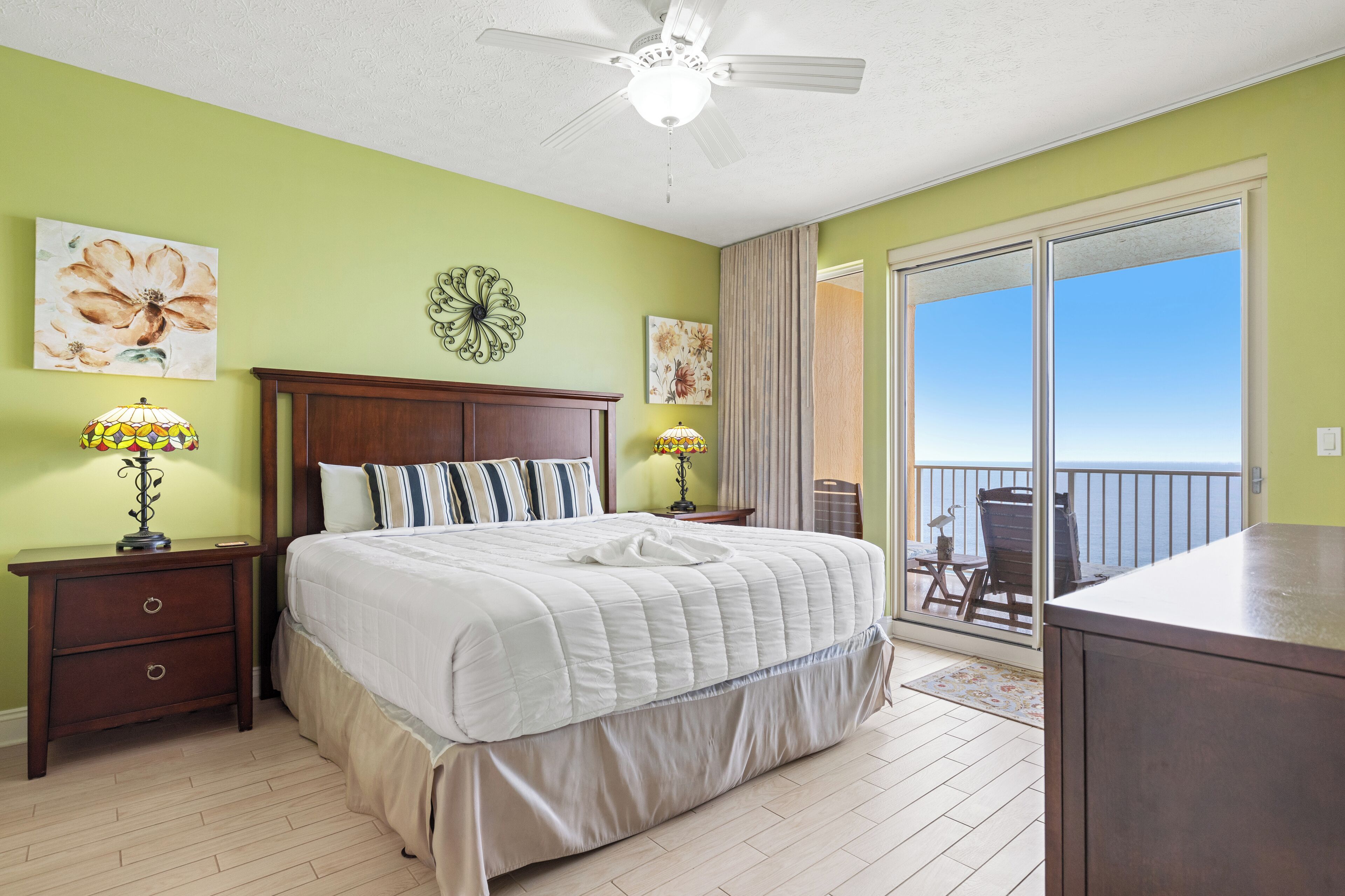 A bedroom with a bed, two nightstands, a ceiling fan, and a balcony with a view.