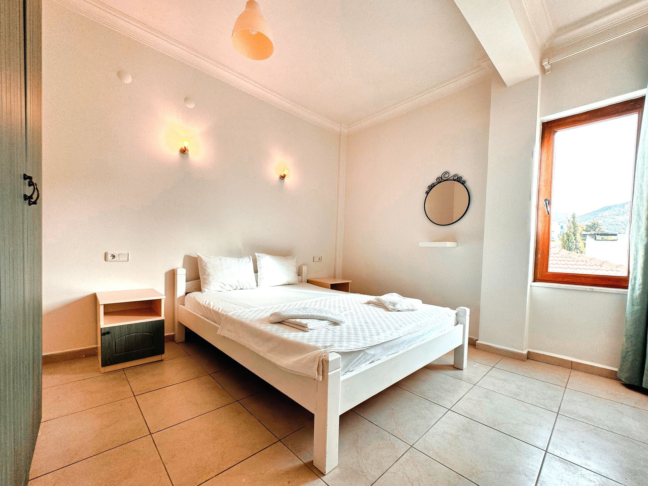 A neatly arranged bedroom with a single bed, bedside tables, a mirror, and a window with a view.