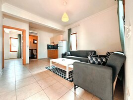 A modern living room with a grey sofa, a white coffee table, and a black and white rug. There is a kitchen area with a refrigerator and a microwave in the background.