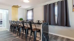 Un comedor con una mesa larga y varias sillas, una planta pequeña en maceta y una ventana con cortinas.