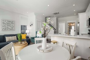 A modern kitchen with white cabinetry, a dining area with a white table and chairs, and a living room with a dark sofa and decorative pillows.