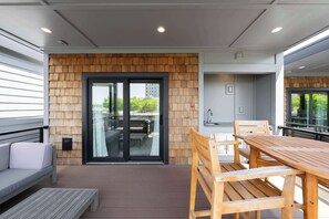 A modern outdoor patio with a wooden deck, a dining table with chairs, a sofa, and a sliding glass door leading to an indoor space.