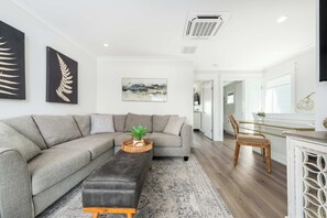 A modern living room with a grey sofa, a black ottoman, and a coffee table with a plant. There is a painting on the wall and a kitchen area visible in the background.
