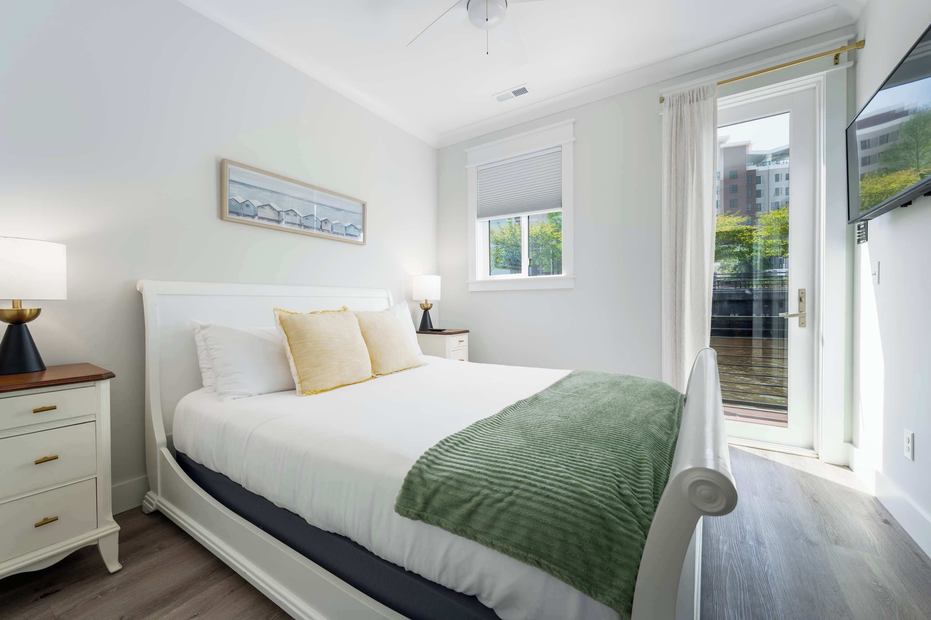 Un dormitorio con cama, mesitas de noche, una ventana con persianas, un cuadro en la pared y vistas al exterior.